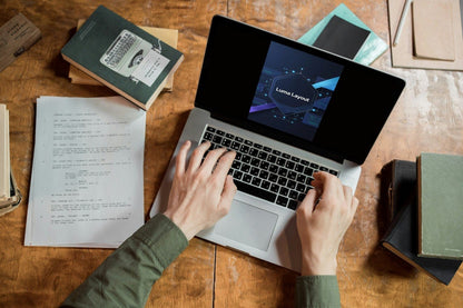Top-down view of a person typing on a silver laptop at a wooden desk, surrounded by books, papers, and notebooks.